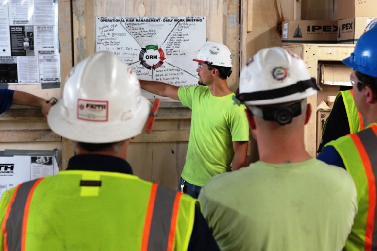 A manufacturing team gathers for a meeting wearing helmets with decals on them